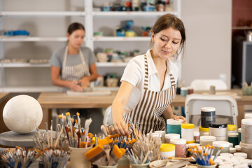 Girl student in pottery workshop stands at table with tools and supplies, picks up brushes for work...