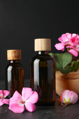 Bottles of geranium essential oil and beautiful flowers on black table, closeup