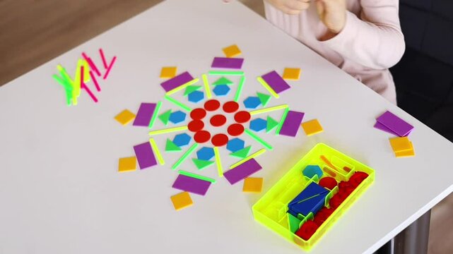 A small European boy assembles a circular mandala pattern from geometric shapes of different colors. 