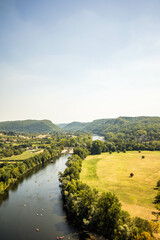 A river winding through a lush green valley, bordered by trees on one side and an open grassy field on the other. Small boats or kayaks are visible on the river under a clear, sunny sky