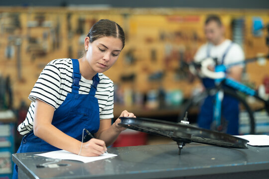 Concentrated skilled young female mechanic attentively examining bike wheel rim and documenting service checklist at counter of bicycle repair workshop
