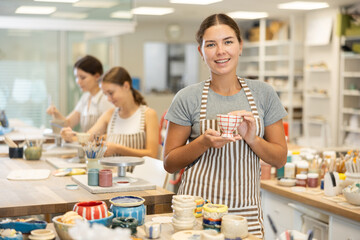 Young female teacher in apron with handmade ceramic cup posing in ceramics workshop