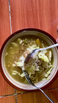 The movement of a spoon and fork stirring Soto Ayam, a typical Indonesian food containing noodles and minced chicken with broth in a bowl on the table.