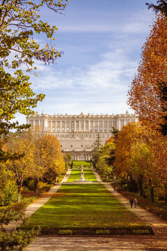 The Royal Palace of Madrid viewed from the Campo del Moro gardens, framed by trees and a clear sky. The palace is in the background, and the garden includes pathways with tourists and a fountain