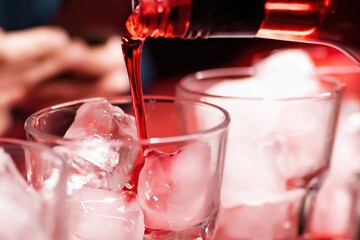 Red syrup is poured from a bottle into a large cocktail glass with ice cubes on the bar counter