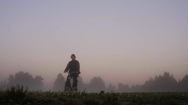 Man Cycling Through Field at Dawn, Passing Camera, Low-Angle Shot, Haze