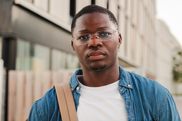 Serious African American student looking pensive at the camera, holding a folder and wearing a backpack, standing outdoors in a modern urban setting, expressing confidence at university campus