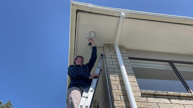 Middle-aged caucasian man standing on a ladder while adjusting an LED security sensor light to the underside of a soffit on the corner of a brick clad 1980s house in New Zealand