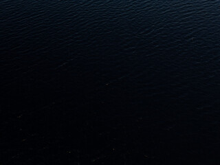 Aerial view of dark, still water captured by a drone, showcasing a serene, almost mysterious atmosphere. The black water surface reflects minimal light, creating a sense of calm and depth.