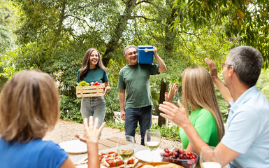 A group of friends gathers in a lush garden, sharing laughter while one person carries fresh vegetables and another holds a cooler. The atmosphere is joyful and inviting, filled with nature.