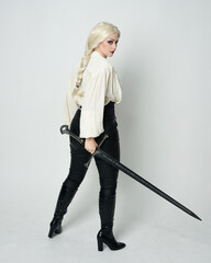 Full length portrait of beautiful female model wearing vintage gothic fantasy blouse, leather pants, boots and blonde plaited hair, larp costume.  standing pose housing sword weapon in fighting action