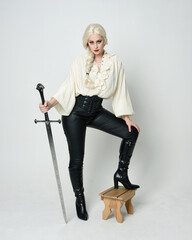 Fototapeta premium Full length portrait of beautiful female model wearing vintage gothic fantasy blouse, leather pants, boots and blonde plaited hair, larp costume. standing pose housing sword weapon in fighting action