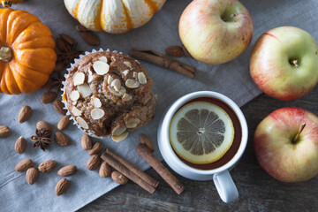 A mug of hot tea with a slice of lemon next to a freshly baked spiced almond and apple muffin on a linen tablecloth on a wooden table decorated with pumpkins and apples