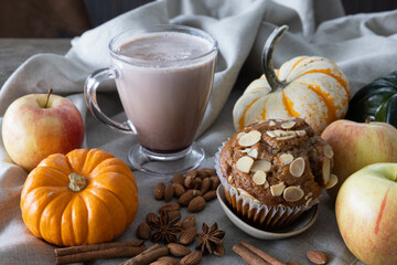 Hot chocolate in a glass mug paired with a freshly baked spiced apple and almond muffin surrounded with pumpkins of various shapes laid on a linen tablecloth