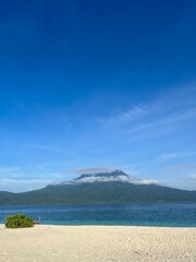 A picturesque beach with pristine white sand, crystal-clear blue water, and a majestic mountain shrouded in mist under a vibrant blue sky.