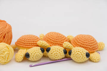 Close up on hand made crochet turtles in yellow and orange on an off white background with crochet hook and balls of yarn on the side.