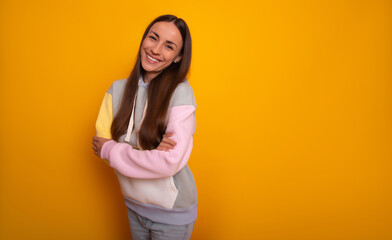 Obraz premium Beautiful smiling young woman stands against a bright yellow background, wearing a pastel hoodie with crossed arms. Her relaxed and cheerful expression exudes a casual, modern, and happy vibe.