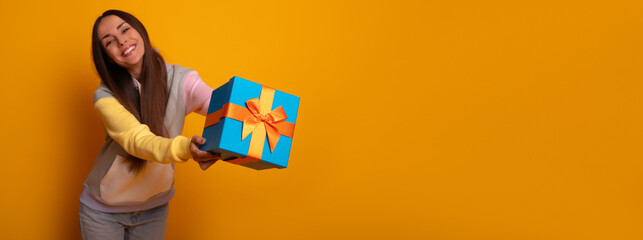 Cheerful young woman extends blue gift box to the camera against vibrant yellow background. Dressed in colorful hoodie, she radiates positivity and generosity. Perfect for holiday or gifting visuals