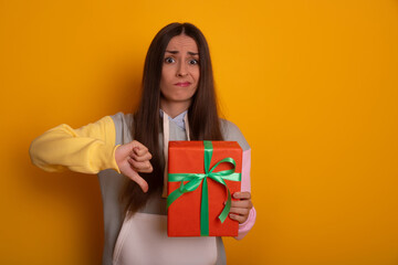 Young woman in hoodie holds red gift box making disappointed expression and giving a thumbs down....