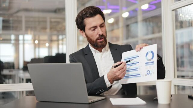 Businessman in a suit sits at his desk in office and reviews charts, graphs of company data on a video conference call. Young worker explaining financial charts on paper slide during online web call.