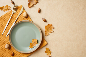 Autumn table setting with a pastel green plate, gold cutlery, and scattered leaves on a textured beige background