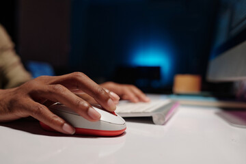 Hand of African American person using computer mouse at desk in dimly lit room with blue lighting in background creating focused and modern ambiance