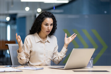 Hispanic woman in office showing frustration during a video call using laptop. Business professional experiencing challenge in work environment. Emotion and remote work concepts included.