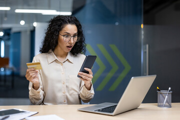 Obraz premium Hispanic woman in office surprised by online transaction, holding credit card, smartphone. Seated at desk, reflecting modern business or shopping.