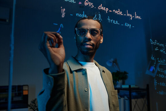Man demonstrated coding on transparent board with floating text around him, wearing glasses and focusing on the task. Room is dimly lit with technology devices in the background