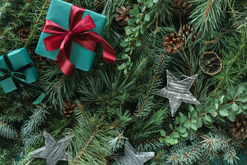 Wrapped gift boxes tied with ribbons on a background of assorted coniferous branches decorated with festive Christmas ornaments.