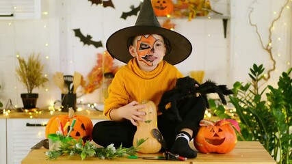 Halloween, a child boy in a festive costume and makeup is preparing for Halloween at home in the kitchen by coloring and carving pumpkins and scaring everyone