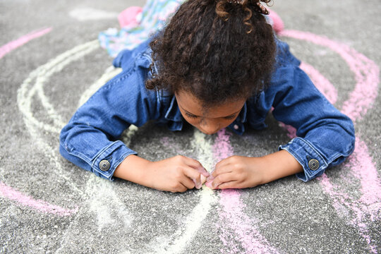 Child lying on pavement, focused on drawing with chalk, denim ja