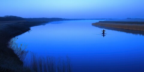 Fototapeta premium A serene blue twilight scene of a person wading through a calm river surrounded by reeds.