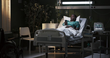 African female patient is lying in the hospital in the ward, holding the phone in her hands, sitting on the Internet, looking at new photos of friends.