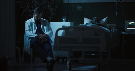 Naklejka premium Doctor sits on a chair next to a seriously ill African woman in a coma, waiting for changes in her condition and puts on stethoscope.
