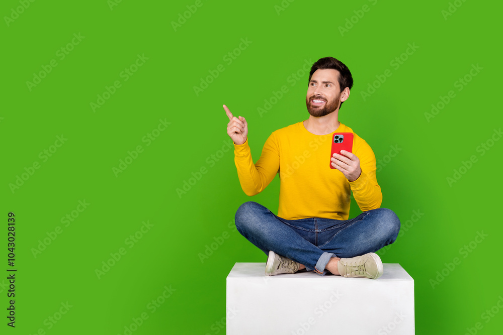 Poster Full body photo of attractive man sit cube point look empty space hold device dressed yellow clothes isolated on green color background
