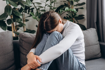 Unhappy anxiety young woman covering face with knee sitting alone on couch in living room at home, hurt heartbroken female in depression feel desperate and anxious having psychological troubles