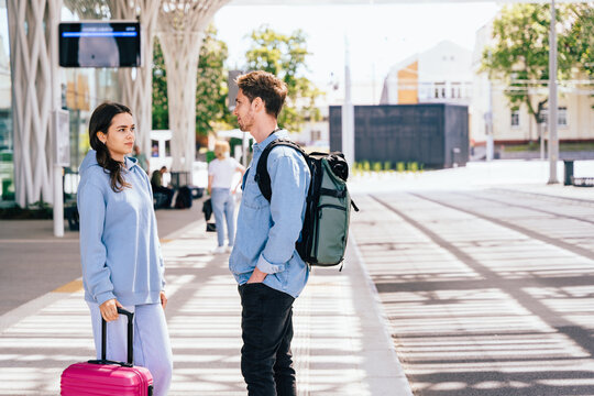 Two young adults, a man and a woman, meeting at modern outdoor transportation hub. Woman holds pink suitcase, while man carries backpack. Bright sunny day enhances atmosphere of travel