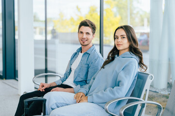 Obraz premium Young Caucasian man and woman sitting in modern waiting area with large windows. Couple travelers casually dressed in relaxed, comfortable clothing. Travel, leisure, and lifestyle concept.