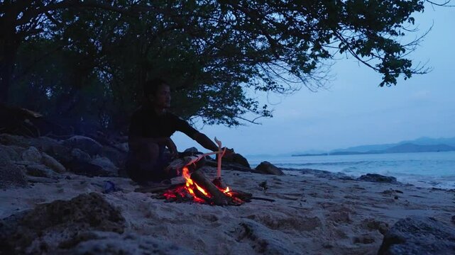 An Indonesian man sitting next to a wood burning fire on the beach cooking a fish on a stick with the ocean and mountains in the background