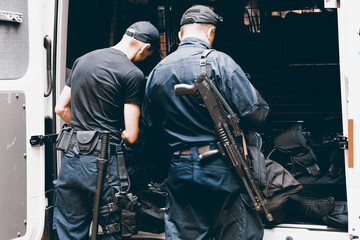 Two armed policemen, from the back. They put on equipment from a police truck. Image filtered