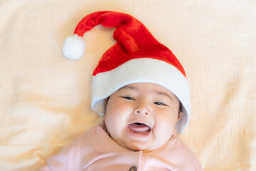 Slant-eyed Caucasian baby girl laughing happily celebrating Christmas in red Santa hat lying on peach colored blanket with copy space