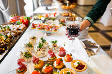 An elegant banquet table with assorted appetizers, including cheese, fruits, and pastries, is set for a celebration. A hand holding a glass of red wine adds a touch of sophistication to the scene