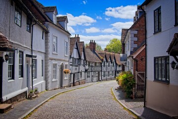  a quaint, cobblestone street lined with traditional, old-fashioned buildings under a clear blue sky. The scene exudes charm and historical ambiance, suggesting a picturesque village