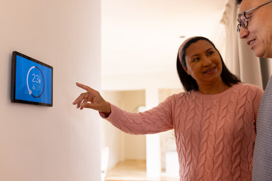 Multiracial senior couple adjusting smart thermostat at home, smiling and discussing settings - Powered by Adobe