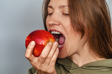 Woman bites red apple, showing teeth. Brown hair. Apple is held in right hand on gray background.