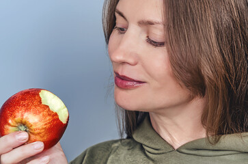 Close-up bitten red apple held by smiling woman. Concept of healthy food and diet. Gray background.
