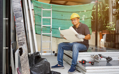Fototapeta premium A smiling man worker in a warehouse holding a clipboard, wearing a hard hat, surrounded by construction equipment and building materials