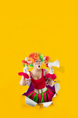 Funny female clown shouting with loudspeaker pointing finger up, looking through the ripped paper hole. Woman entertainer as colorful Joker, suit and wig, clown whiteface makeup, professional actor.