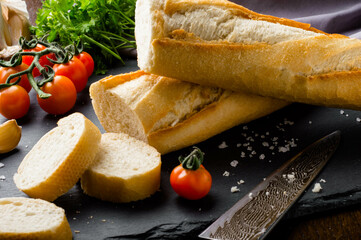 Fresh bread on black stone background. Fresh crispy french baguette on on a slate board, bakery products. Rustic bread slices with fresh tomatoes and parsley.